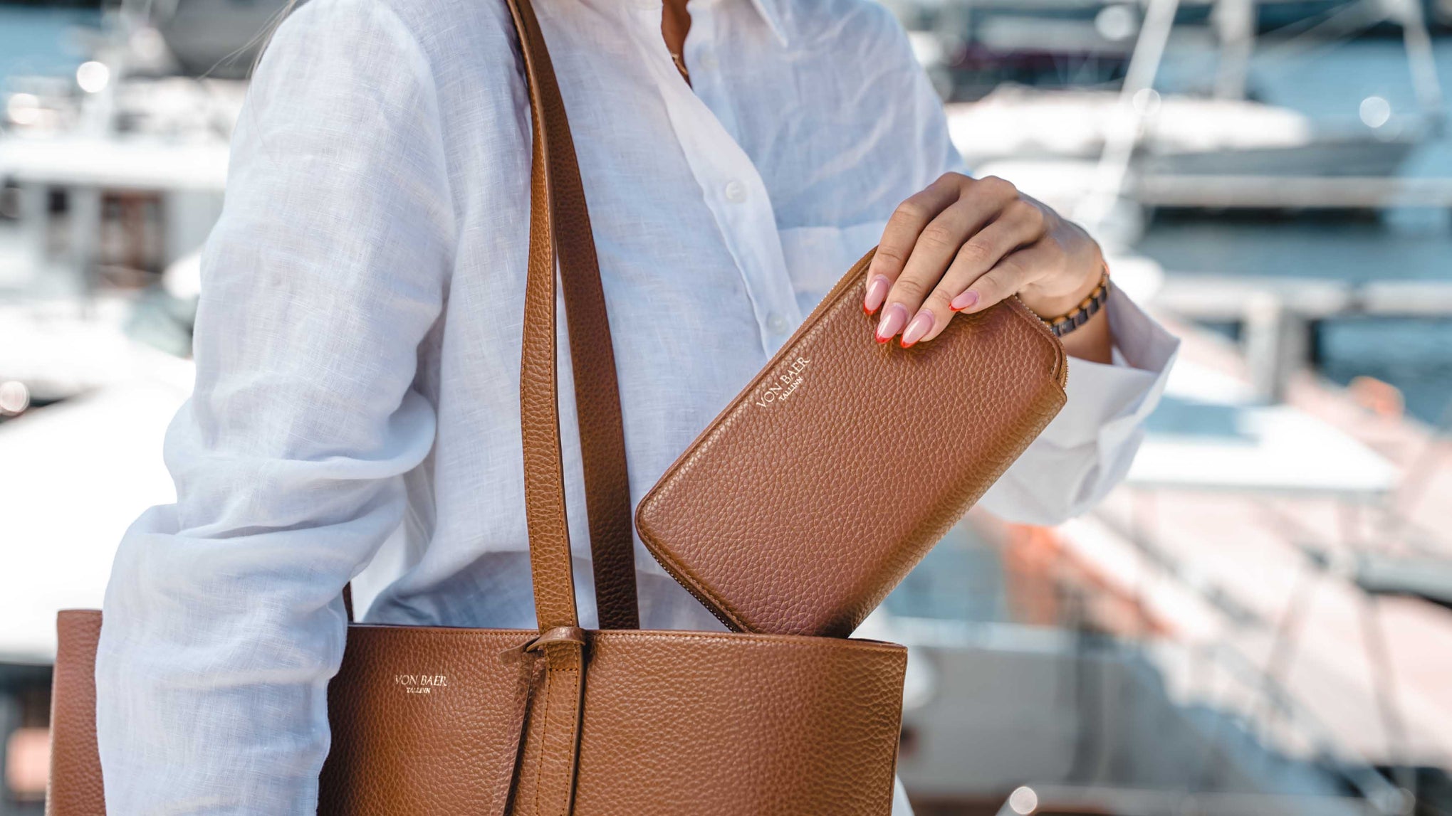 A person in a white linen shirt carries a brown pebbled leather tote bag and holds a matching brown leather zip-around wallet, with a blurred marina background.