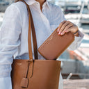 A person in a white linen shirt carries an amazing looking vb tan pebbled leather tote bag for women and holds a matching tan leather wallet. The background shows a blurred outdoor marina setting with boats.