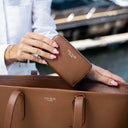 A person holds a good looking tan pebbled leather zip-around wallet with gold-toned VON BAER lettering, resting it on top of a matching brown leather tote bag in an outdoor setting.