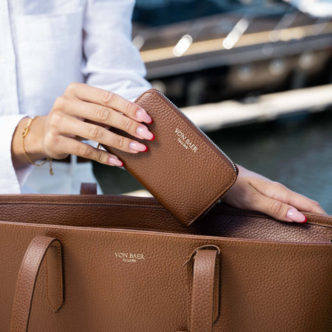 A person holds a good looking tan pebbled leather zip-around wallet with gold-toned VON BAER lettering, resting it on top of a matching brown leather tote bag in an outdoor setting.