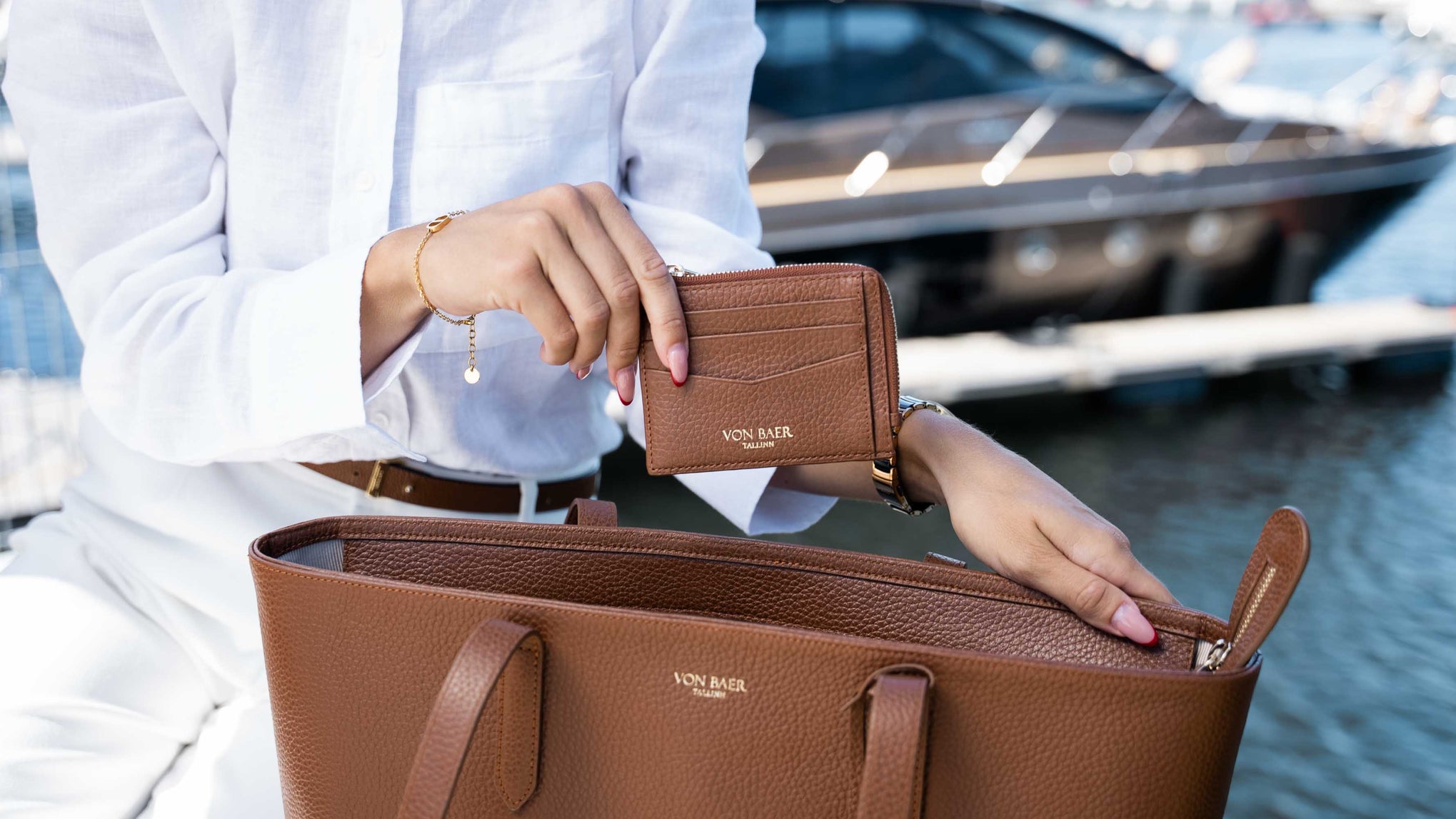 A person in a white outfit holds a brown pebbled leather wallet branded 'Von Baer' while placing it into a matching brown tote bag, with a blurred marina and yacht in the background.