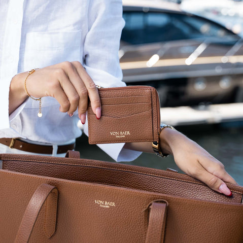 A person in a white shirt holds a brown leather card holder wallet branded 'VON BAER' next to a matching brown pebbled leather tote bag, with a blurred yacht in the background.