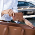 A person in a white shirt holds a brown leather card holder wallet branded 'VON BAER' next to a matching brown pebbled leather tote bag, with a blurred yacht in the background.