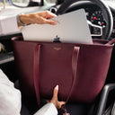 A burgundy pebbled leather tote bag with gold 'VON BAER TALLINN' lettering, held inside a porsche car by a person whose hands and arms are visible, with a silver laptop partially inserted into the top.