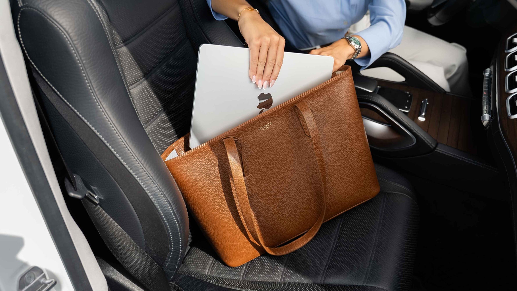 A brown leather von baer tote bag with a silver laptop macbook inside a black car porsche seat. A person wearing a light blue shirt is partially visible, holding the laptop in the bag.