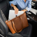 A woman in a light blue shirt sits in a black car seat and places a silver laptop into a brown pebbled von baer leather tote bag. The bag has matching leather handles and a small gold logo on the front.