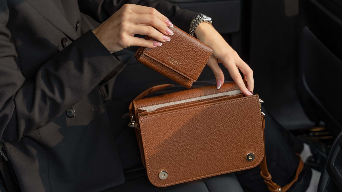 Von Baer Elise burgundy trifold wallet for women shown alongside an Estelle small leather tote bag, held by a person in a black blazer inside a car interior for a luxury handbag detail view.
