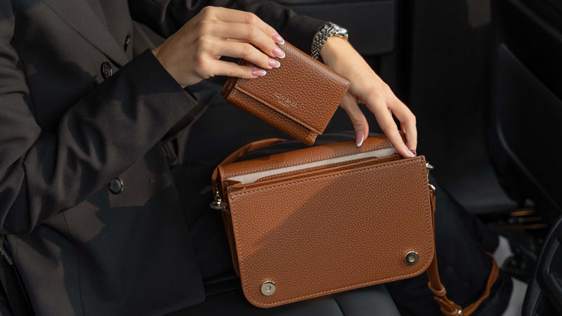Von Baer Elise burgundy trifold wallet for women shown alongside an Estelle small leather tote bag, held by a person in a black blazer inside a car interior for a luxury handbag detail view.