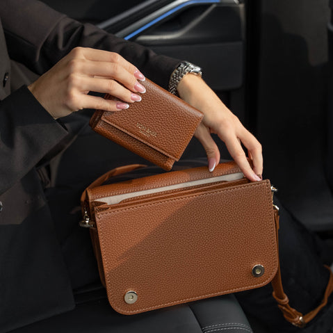 Von Baer Elise trifold wallet and Estelle small tote bag in burgundy pebbled leather shown as a person in a black blazer holds the wallet above the matching bag inside a car.