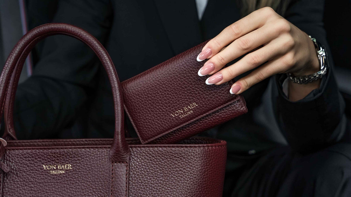 Von Baer Elise burgundy trifold wallet for women shown over a matching Estelle small leather tote bag, held by a person wearing a black blazer and silver watch inside a luxury car interior.