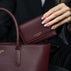 Von Baer Elise trifold wallet and Estelle small tote bag in burgundy pebbled leather held together in a luxury car setting, showing gold foil brand lettering.
