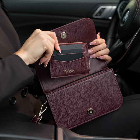 Von Baer Isabella small burgundy pebbled leather crossbody bag shown with the Essence women's card case fitting inside, held in a car interior and featuring embossed Von Baer Tallinn branding.