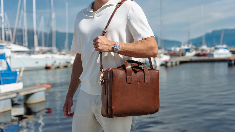 A man in a white polo shirt and trousers carries a brown luxury full grain leather briefcase-style bag from von baer over his shoulder while walking near a marina with boats and water in the background.