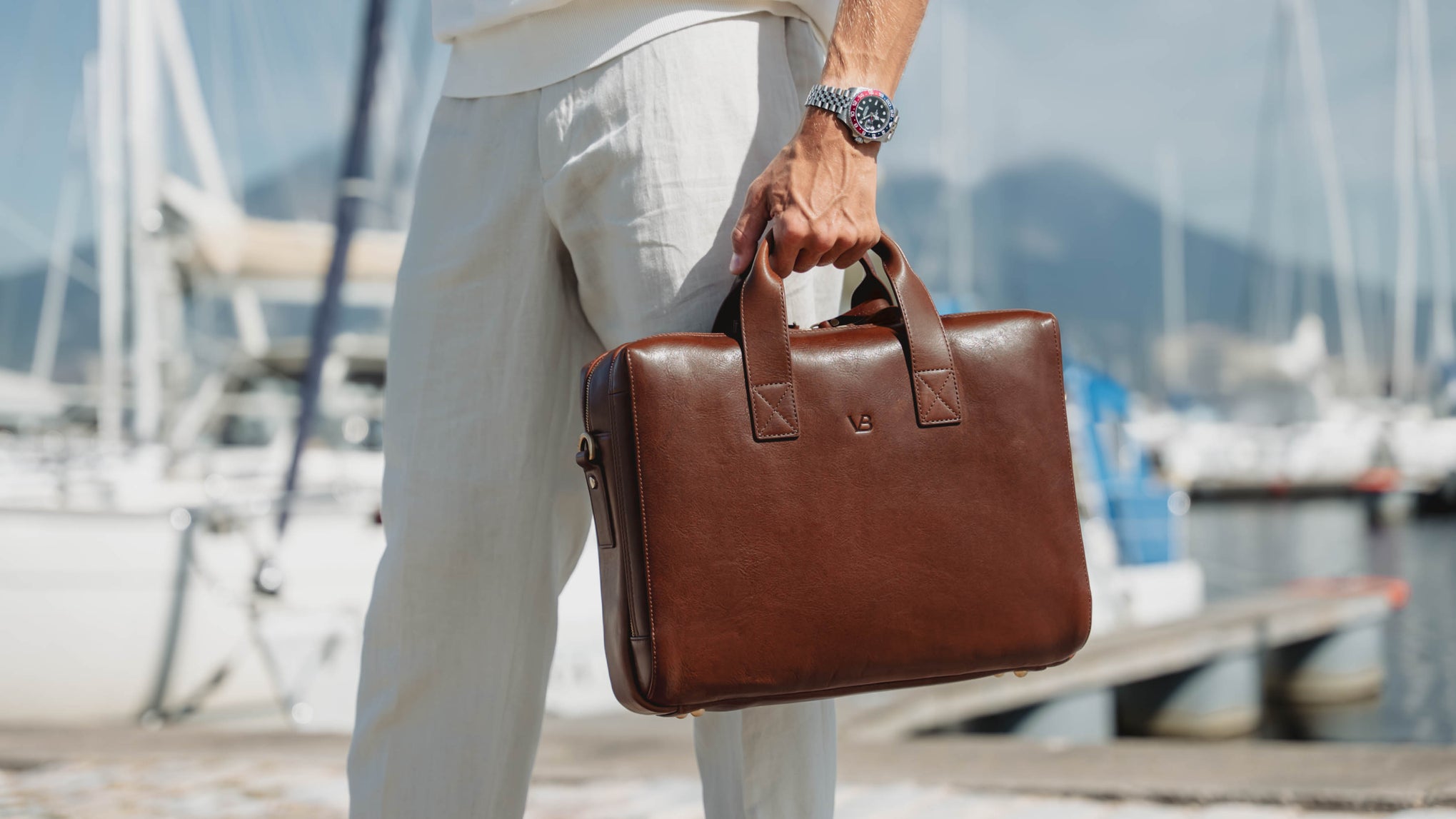A man in white linen clothing holds a brown full grain luxury leather briefcase with a small vb logo, set against a blurred background of a marina with boats and water.