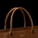 Close-up of a brown pebbled leather handbag with double top handles and a small gold-toned VON BAER logo on the front, set against a solid black background.