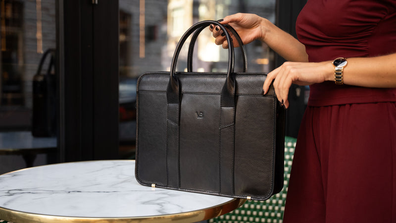 A female model in a red suit is shown with a black Von Baer exquisite luxury leather laptop bag for women, which is placed on a marble table.