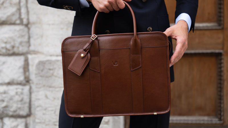 An exquisite, slim, brown Von Baer full-grain leather laptop briefcase with a luggage tag, held by a businessman