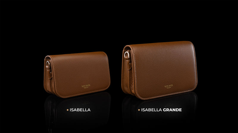 Von Baer Isabella and Isabella Grande women's full-grain pebbled leather crossbody bags in cognac and tan, displayed side by side on a reflective black background to compare sizes, with the smaller Isabella bag on the left and the larger Isabella Grande bag on the right