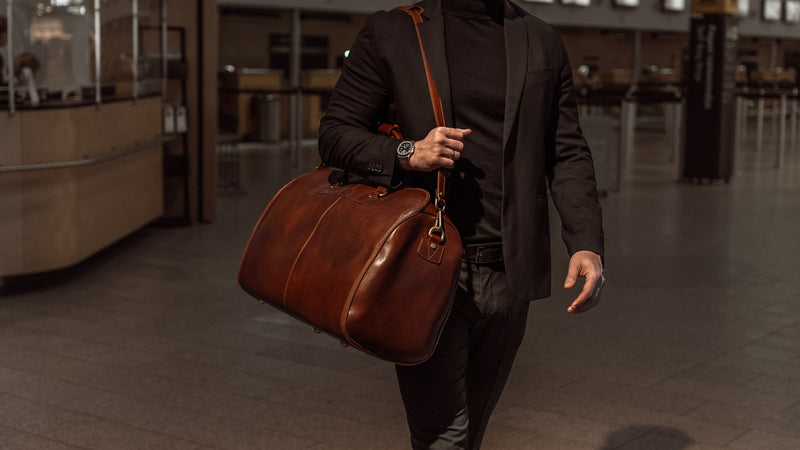 A male model in an airport is carrying a brown Von Baer large luxury leather garment bag with a shoulder strap.