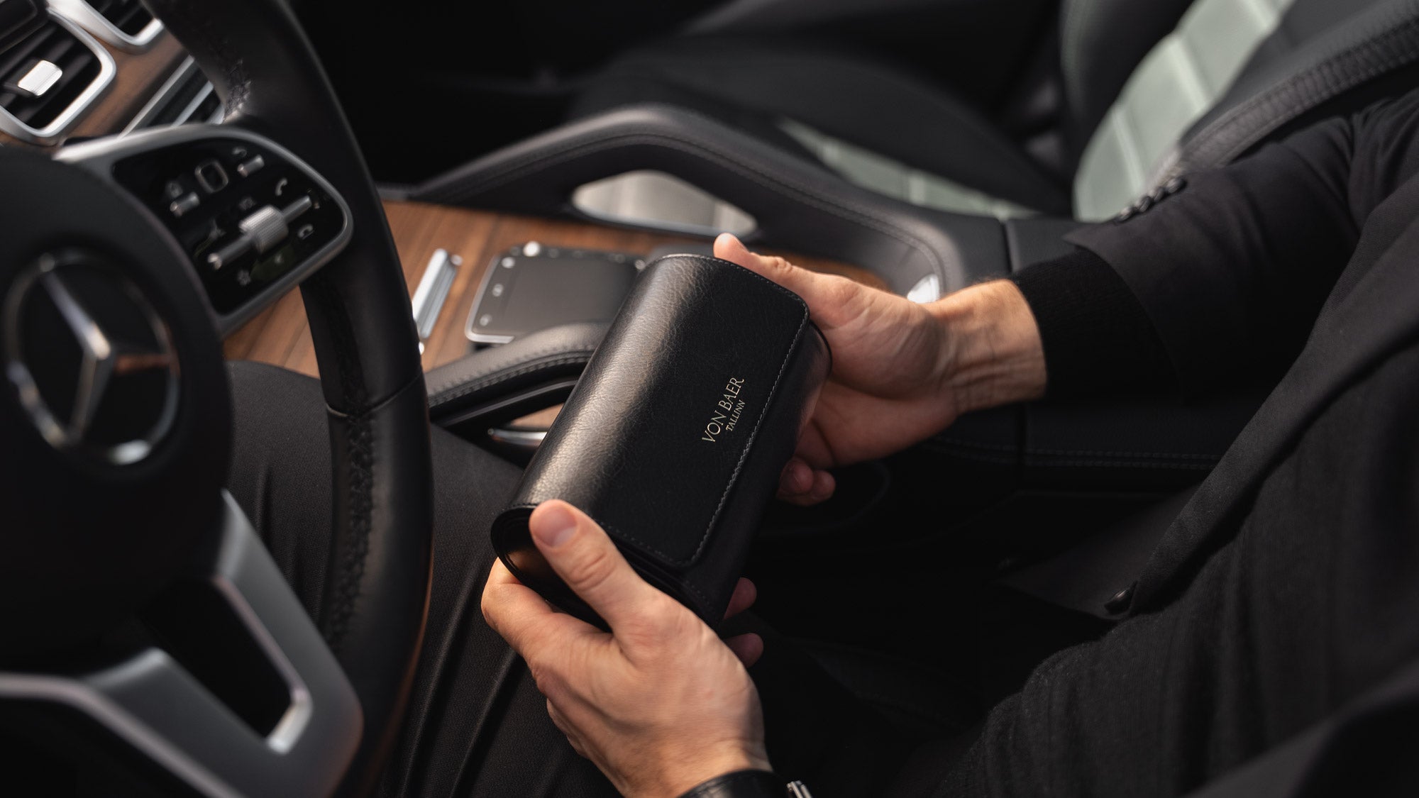A man in a Mercedes car holding a black Von Baer luxury full-grain leather watch roll case with two slots.