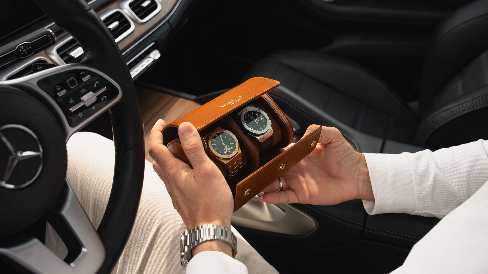 A man in a Mercedes car holding an opened tan Von Baer luxury full-grain leather watch roll case with two slots, showing watches inside.