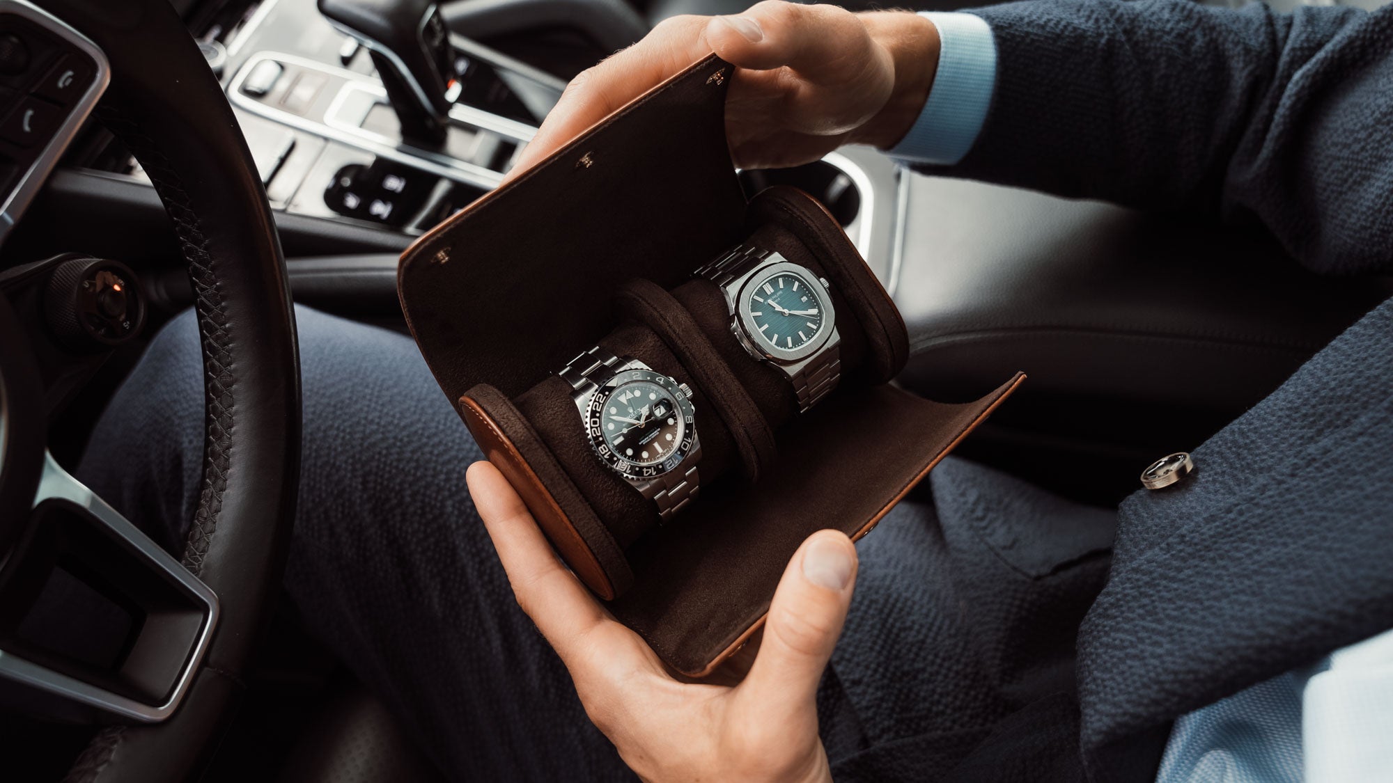 Person holding a luxury brown leather watch box with two watches inside, a Rolex and a Patek Philippe, while sitting in a car.