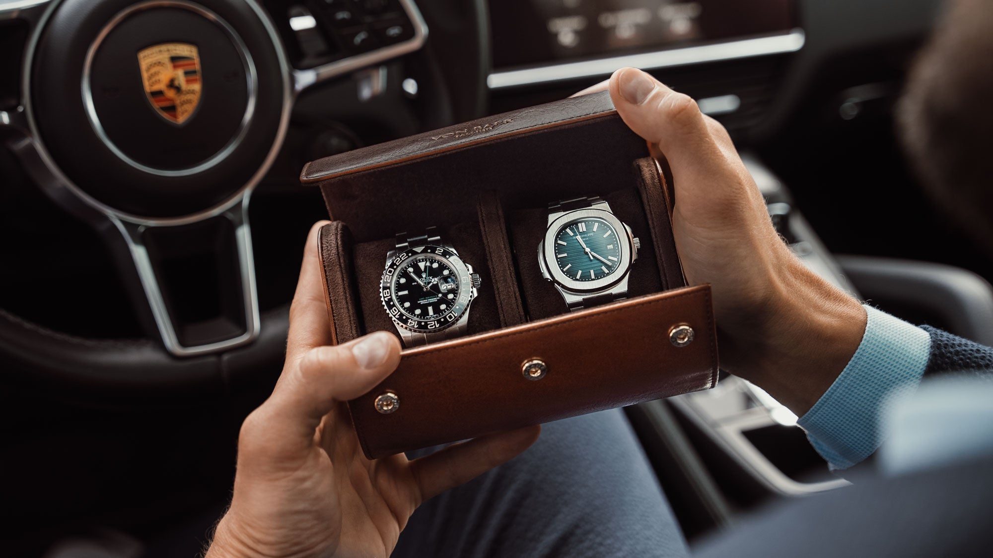 A brown Von Baer luxury leather watch roll with a Rolex and a Patek Philippe watch inside, sitting in a Porsche car.