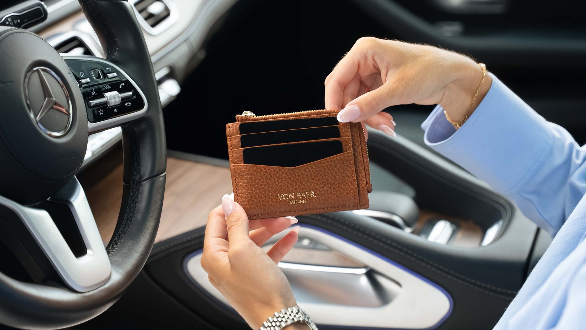 A person holding a brown textured leather card holder wallet with 'Von Baer' embossed on the front, set against a car interior background with a Mercedes-Benz steering wheel visible.