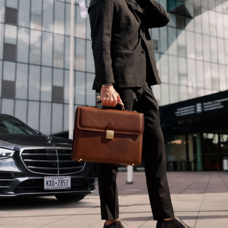 Person in a suit holding a brown leather briefcase in front of a modern building with a car in the background.