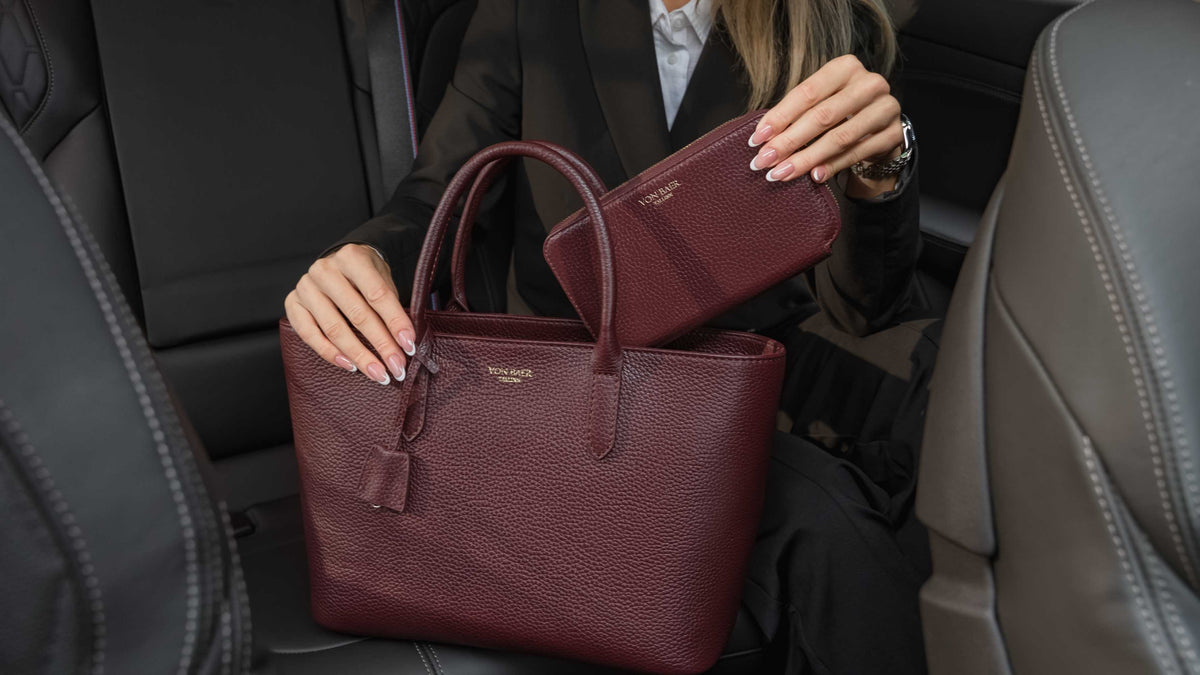 Von Baer Origin Portmonnaie burgundy luxury leather wallet shown alongside an Estelle small leather tote bag, held by a person inside a car interior for a premium lifestyle view.