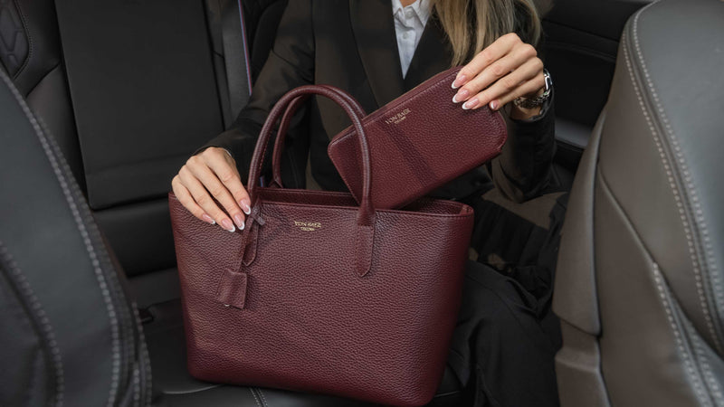 Von Baer Origin Portmonnaie burgundy luxury leather wallet shown alongside an Estelle small leather tote bag, held by a person inside a car interior for a premium lifestyle view.
