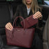 Von Baer Origin Portmonnaie burgundy luxury leather wallet shown alongside an Estelle small leather tote bag, held by a woman in a black blazer inside a car interior for a premium lifestyle view.