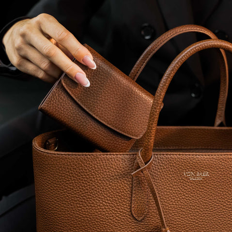 Von Baer Reverie compact women's pebbled leather wallet in cognac, being placed into an Estelle small leather tote bag with double top handles and a gold-tone logo plaque, shown in a close-up against a dark background