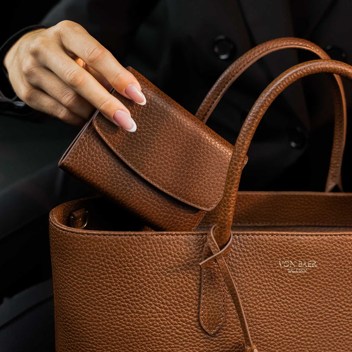 Von Baer Reverie compact women's pebbled leather wallet in cognac, being placed into an Estelle small leather tote bag with double top handles and a gold-tone logo plaque, shown in a close-up against a dark background