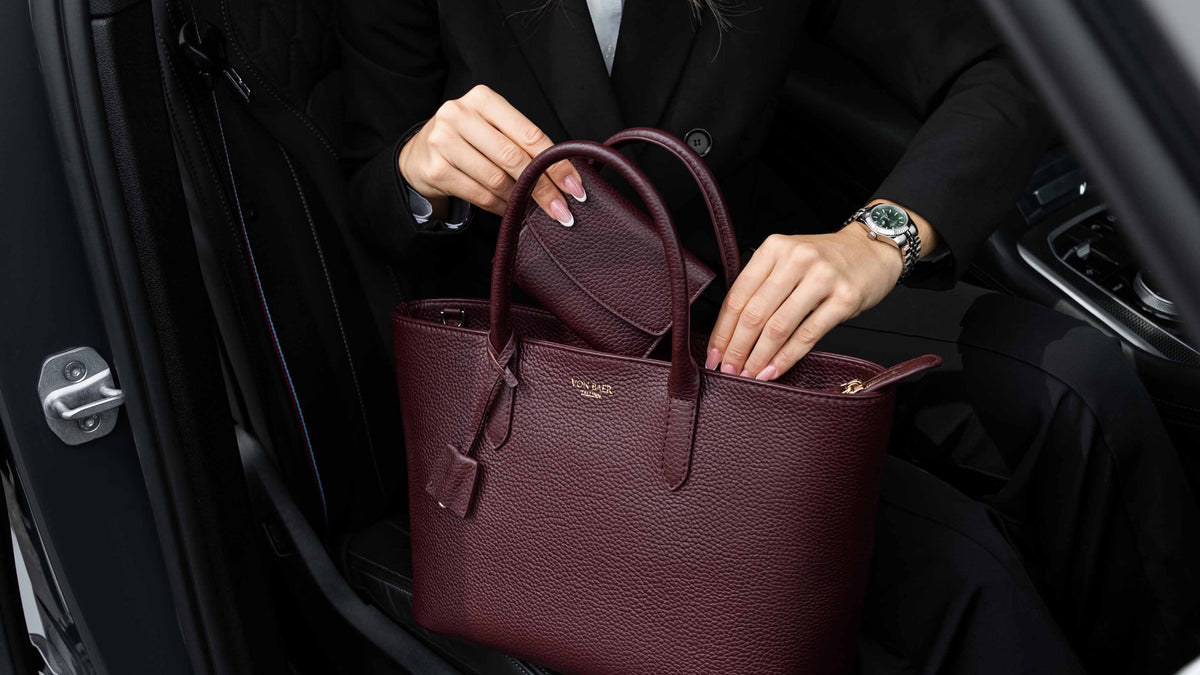 Von Baer Reverie burgundy women's full-grain leather wallet shown alongside an Estelle small leather tote bag, held by a person in a black blazer inside a car interior for a lifestyle view.
