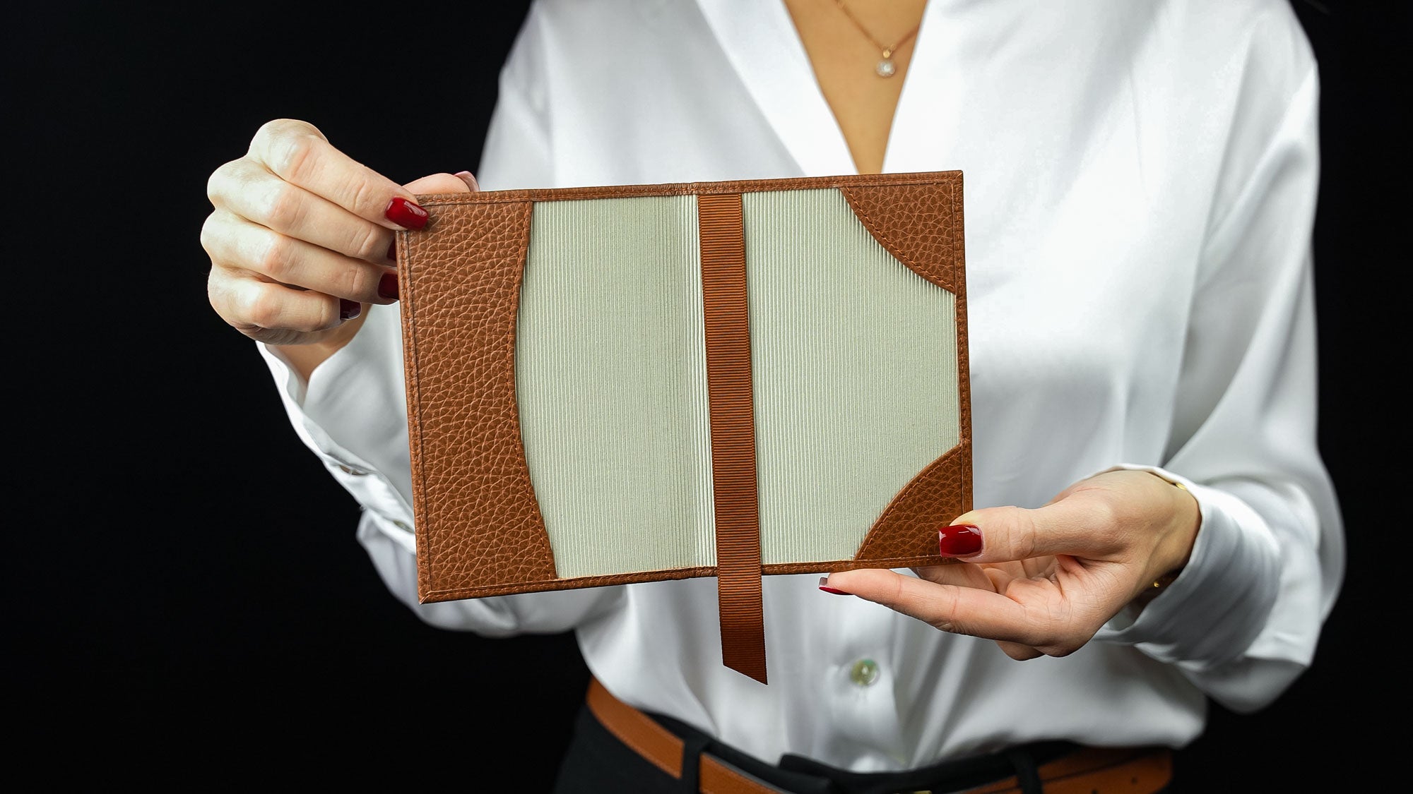 Person holding a leather passport holder with a brown strap against a black background