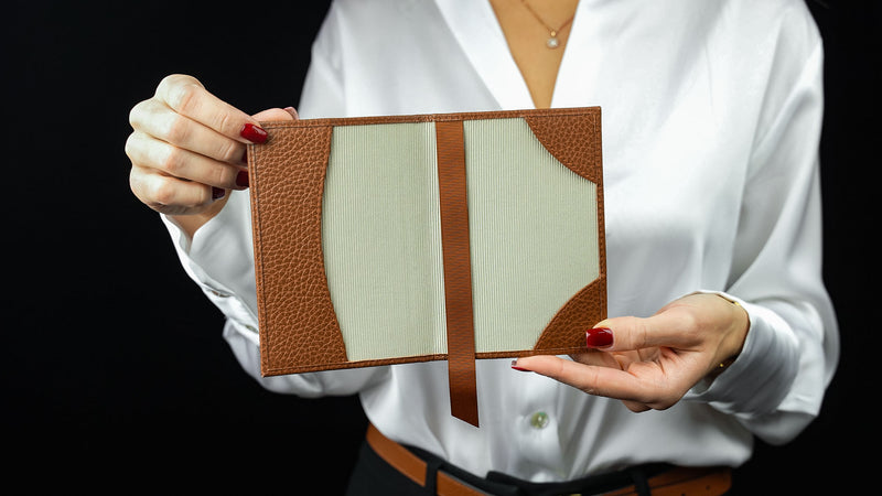 Person holding a leather passport holder with a brown strap against a black background