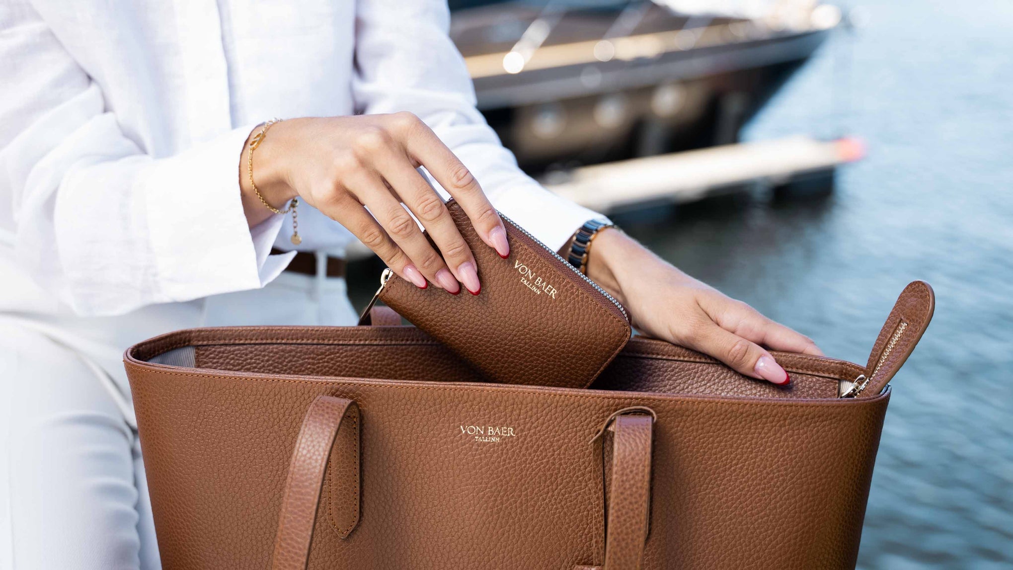 A person in a white outfit organizes a classic tan pebbled leather tote bag and matching zip wallet on a dock with water and a boat in the background. The brand name 'VON BAER' is visible on both items.