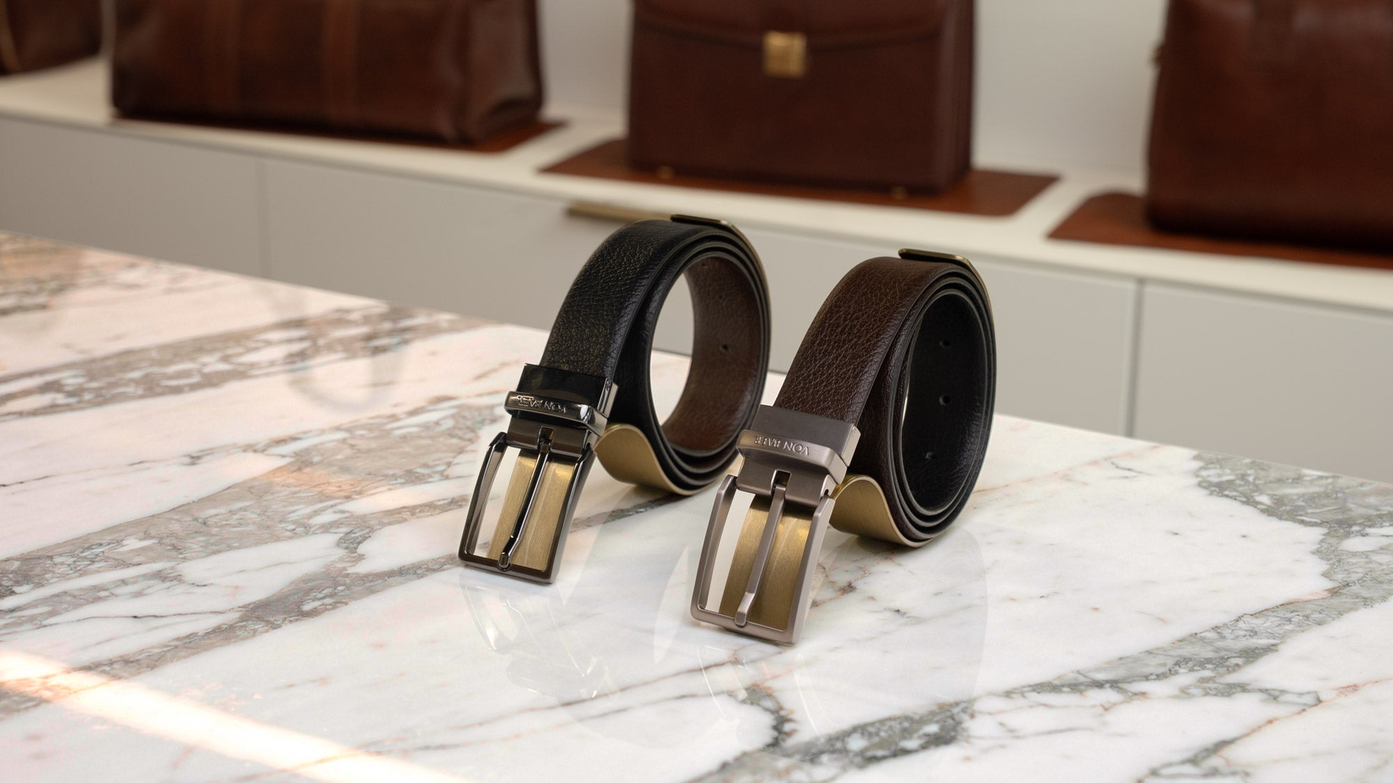 Von Baer Premier full-grain 2-in-1 reversible leather belt in brown and black, with black and brushed graphite buckles displayed side by side on a luxury marble table in a boutique setting