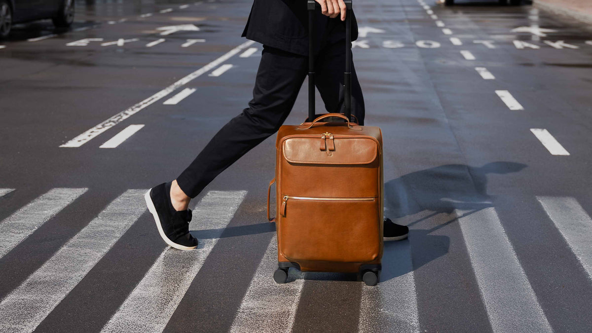 Von Baer Voyager luxury full-grain leather carry-on trolley bag in brown, pulled by a stylish man dressed in black across a wet city crosswalk, showcasing its elegant design and travel functionality