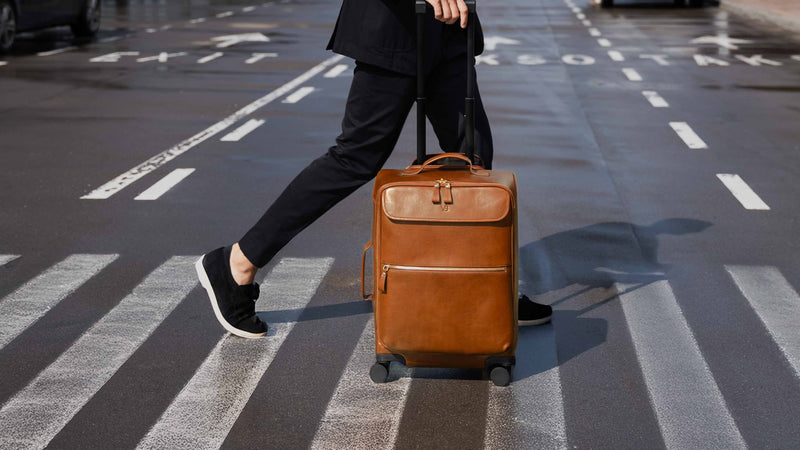 Von Baer Voyager luxury full-grain leather carry-on trolley bag in brown, pulled by a stylish man dressed in black across a wet city crosswalk, showcasing its elegant design and travel functionality