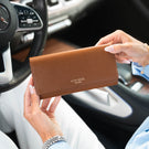 A person holding a tan pebbled leather purse with 'VON BAEr' printed on the front, set against a blurred car interior background.