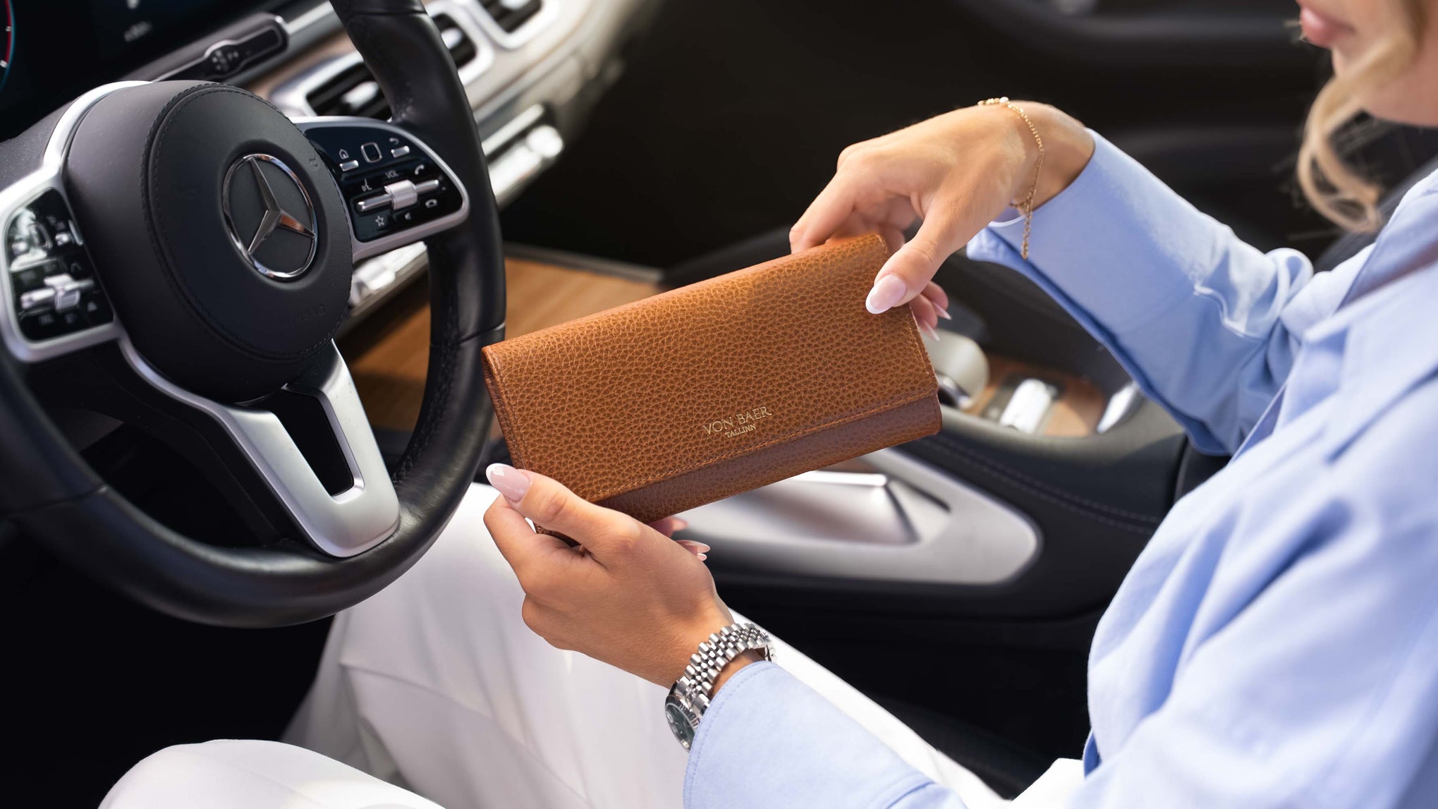 A woman in a light blue shirt holds a vb cognac tan textured leather wallet inside a Mercedes-Benz car, with the steering wheel and dashboard visible.