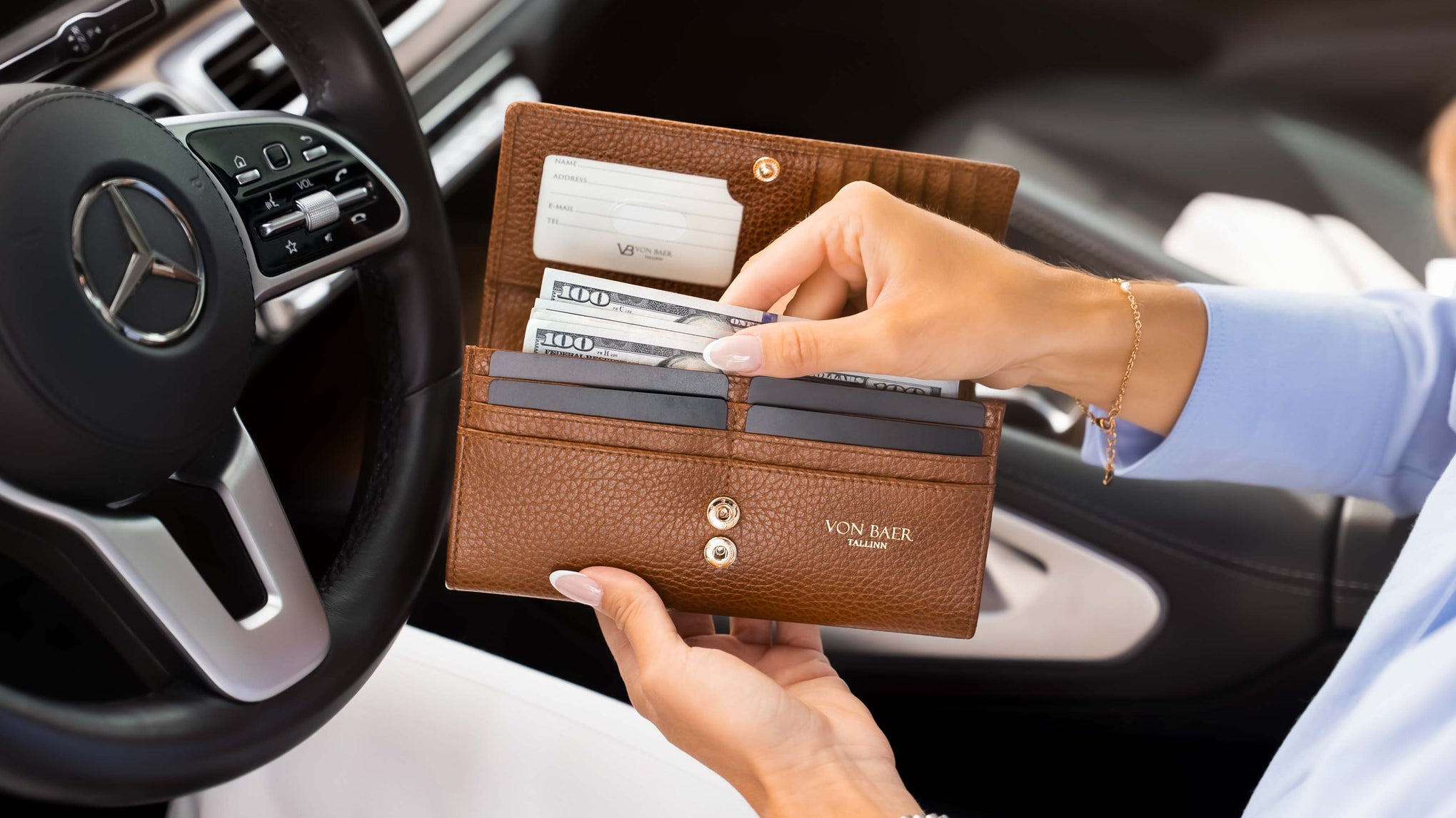 A brown pebbled luxurious tan leather wallet by Von Baer, held open by a person inside a car. The wallet contains credit cards and cash, with the Mercedes-Benz steering wheel visible in the background.