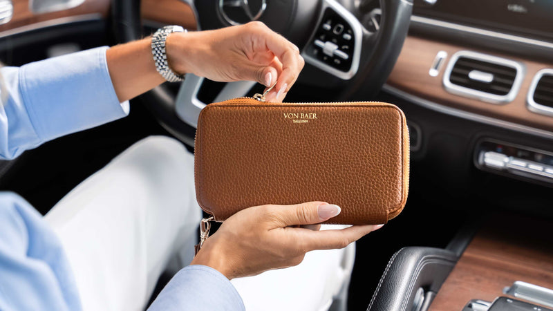 A woman in a light blue shirt holds a tan textured leather zip-around wallet with gold lettering that reads 'VON BAER' inside a luxurious car cabin with wood trim.