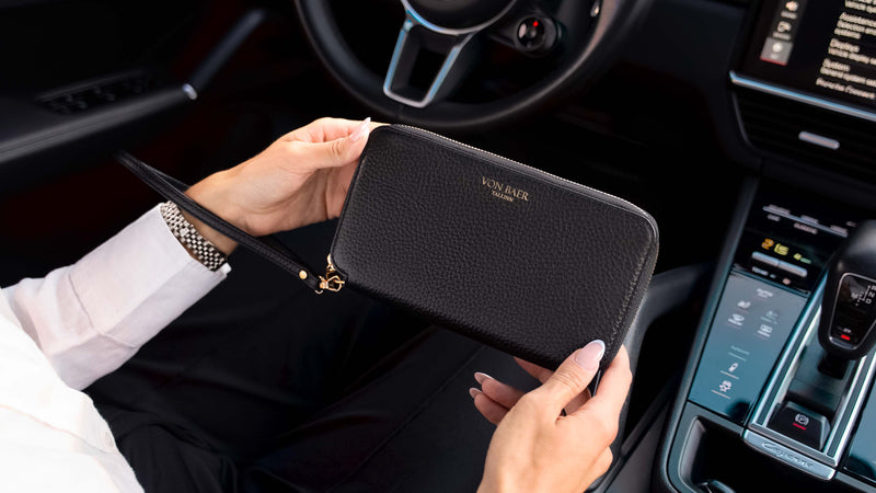 A person holding a nice looking black pebbled premium leather zip-around wallet with gold hardware inside a car cabin. The wallet features the brand name 'Von Baer' in gold lettering on the front.