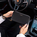 A person holding a black pebbled leather Von Baer zip-around wallet inside a porsche car, with the steering wheel and dashboard visible in the background.
