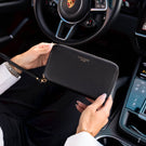 A person holding a black pebbled leather Von Baer zip-around wallet inside a porsche car, with the steering wheel and dashboard visible in the background.