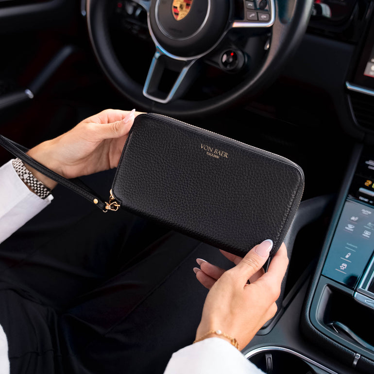 A person holding a black pebbled leather Von Baer zip-around wallet inside a porsche car, with the steering wheel and dashboard visible in the background.