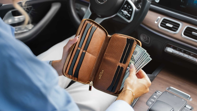 A person sitting in a car holds an open tan leather wallet showing card slots and cash compartments, with hands and part of the car interior visible.